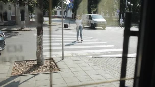 Minsk, Biélorussie 20 septembre 2020 : Une jolie jeune fille en T-shirt blanc et en jean traverse la rue à une intersection et entre dans le bâtiment par une porte vitrée. Vue de face. Tir de l'intérieur — Video