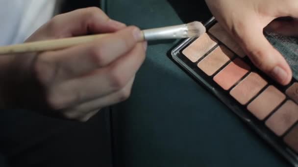 Una joven usa una paleta de sombras de ojos pastel para maquillarse y lleva un cepillo cosmético en la mano. Primer plano de las manos — Vídeos de Stock