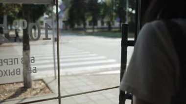 Beyaz tişörtlü ve kot pantolonlu genç bir kadın camdan bir kapıdan dışarı çıkar ve yolun karşısına geçer. Arkadan bak. Binanın içinden ateş ediliyor.