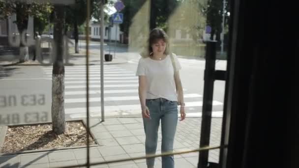 Una joven atractiva camina hacia el edificio y atraviesa la puerta de cristal. Vista frontal. Disparos desde el interior del edificio — Vídeo de stock
