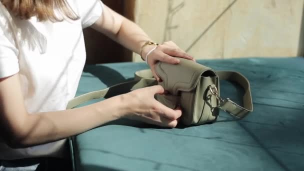 Una joven con una camiseta blanca se sienta en una mesa frente a la ventana y sacude varios cosméticos de su bolso. Primer plano de las manos — Vídeos de Stock