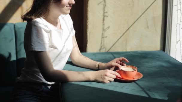 Beyaz tişörtlü şirin bir kız bir kahve fincanı ve gülümsemesiyle bir şehir kafesinin penceresinin yanında oturuyor. Yan görüş. Yakın plan. — Stok video