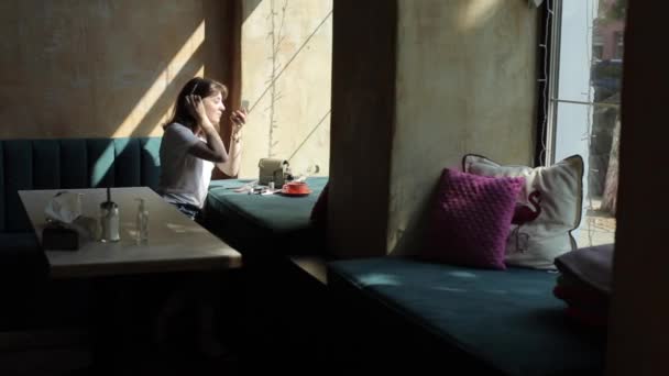 Una linda joven con una camiseta blanca se sienta junto a la ventana en un café y extiende cosméticos en el alféizar de la ventana cerca de una taza de café hace un maquillaje. Plan global — Vídeo de stock
