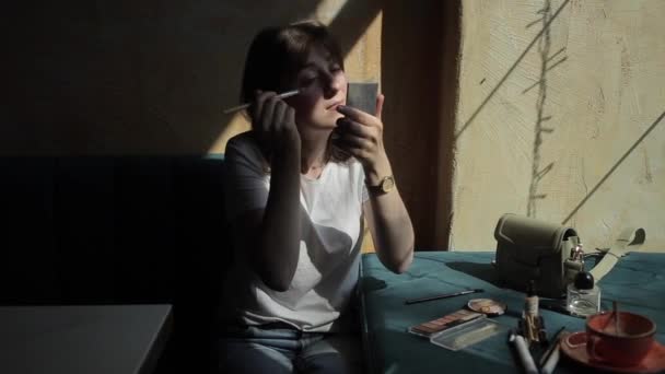 Приємна молода жінка в білій футболці і джинсах сидить в кафе і розкидає косметику на столі робить її макіяж перед дзеркалом. Вид спереду — стокове відео