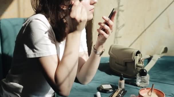 U stolu v kavárně sedí energická mladá dívka v bílém tričku, dívá se do zrcadla a v ruce drží kosmetickou tužku. Boční pohled. Detailní záběr — Stock video