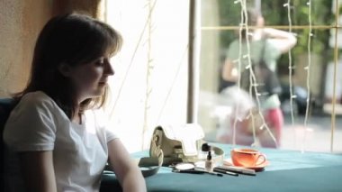 Beyaz tişörtlü neşeli genç bir kadın bir kahve dükkanının penceresinde oturmuş birileriyle konuşuyor ve aynaya bakıp gülüyor. Yan görüş. Yakın plan.