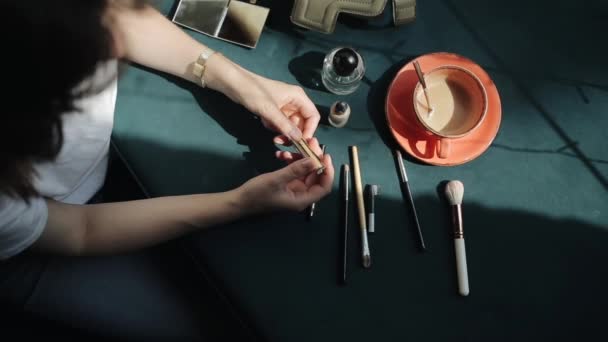Mladá dívka v bílém tričku se chystá udělat make-up a rozložit kosmetiku na stůl vedle šálku kávy. Pohled shora. Detailní záběr — Stock video