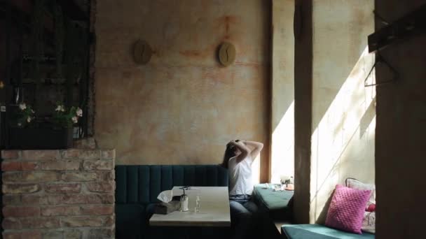 Uma jovem encantadora em uma camiseta branca e jeans se senta em um café na frente da janela e coleta cosméticos em sua bolsa. Vista lateral. Plano global — Vídeo de Stock