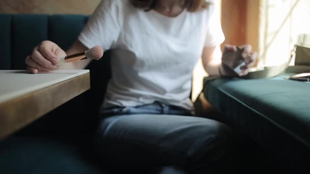 Ein junges Mädchen in weißem T-Shirt und Jeans sitzt in einem Café und klopft mit einem Pinsel auf den Tisch, um überschüssiges Pulver abzuschütteln. Frontansicht. Nahaufnahme. Unklarer Hintergrund — Stockvideo