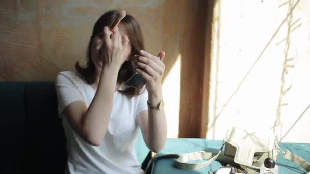 Uma jovem gentil senta-se em um café e faz maquiagem olhando no espelho e aplicando pó em seu rosto com uma escova. Vista frontal. Close-up — Vídeo de Stock