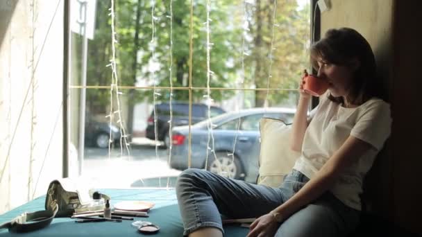 Uma jovem encantadora está sentada no peitoril da janela em um café encostado à parede e bebendo café sorrindo e se alongando com prazer. Vista lateral. Plano médio — Vídeo de Stock