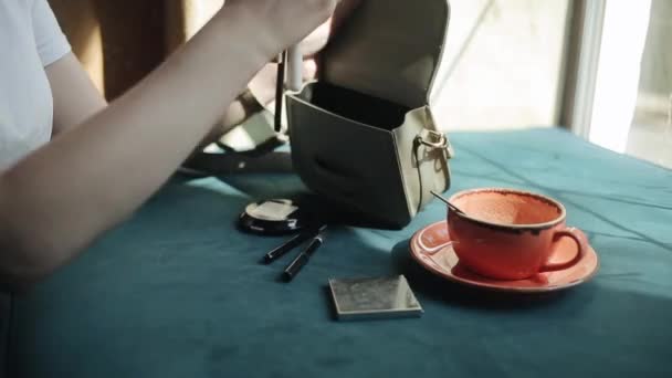 Una joven con una camiseta blanca pone su cosmética fuera de la mesa en su bolso y se va dejando una taza vacía en la mesa. Primer plano. Vista lateral — Vídeos de Stock