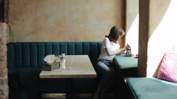 En ung leende kvinna i vit t-shirt och jeans sitter på ett café och efter att ha lagt sina saker i handväskan stiger hon upp från bordet och går. Framifrån. Övergripande plan — Stockvideo