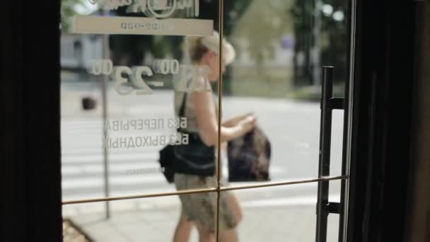 Uma jovem garota em uma camiseta branca deixa um café através de uma porta de vidro em uma rua da cidade em um dia de verão. Vista pelas traseiras. Close-up — Vídeo de Stock