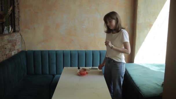 En vakker, ung jente i hvit T-skjorte og jeans lager et oppsett for flate legbilder på en mobiltelefon fra kosmetikk og en kopp mens hun står ved et kafebord. Sett fra siden – stockvideo