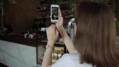 Misk, Belarus - 25 Ağustos 2020: Hoş bir genç kız kafe salonunda duruyor ve cep telefonuyla içerinin fotoğrafını çekiyor. Arkadan bak. Yakın plan. Arka plan bulanık. Telefona odaklan.