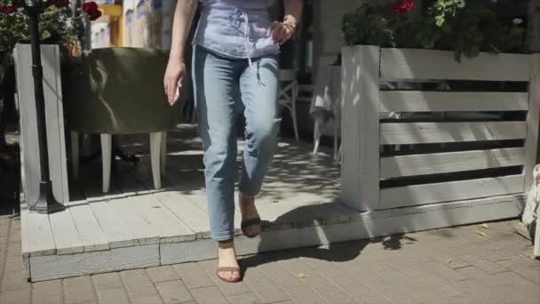 An attractive young woman passes between the tables and sits down on the edge of the wooden platform of an outdoor cafe with her legs stretched out. — Stock Video