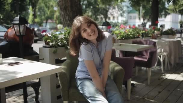 A nice young girl poses in front of the camera while sitting in a chair of a summer cafe against the background of lanterns and flowerpots with flowers. Front view. Medium plan — Stock Video