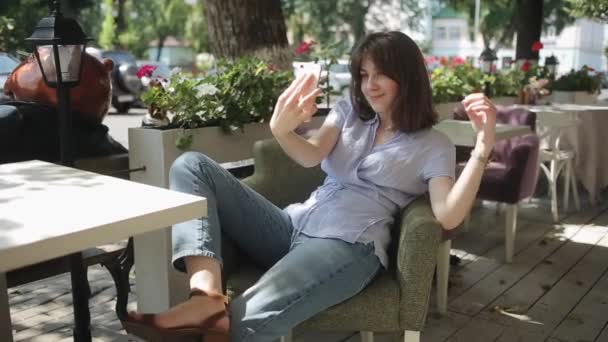 Příjemná mladá dívka v světle modré blůze sedí u stolu v letní kavárně na pozadí městské ulice a bere selfie. Boční pohled. Střední plán. Rozmazané pozadí — Stock video
