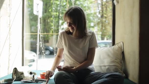 Ніжна молода дівчина сидить на м'якому підвіконні в кафе на фоні вікна і примушує її обличчя сміятися в камері. Вид спереду. Крупним планом — стокове відео