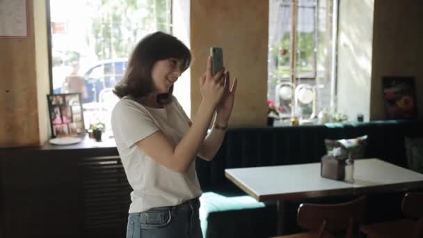 Misk, Weißrussland - 25. August 2020: Eine junge lächelnde Frau in weißem T-Shirt und Jeans steht in einem Café und macht ein Selfie mit einem Handy. Seitenansicht. Mittlerer Schuss. Unklarer Hintergrund — Stockvideo