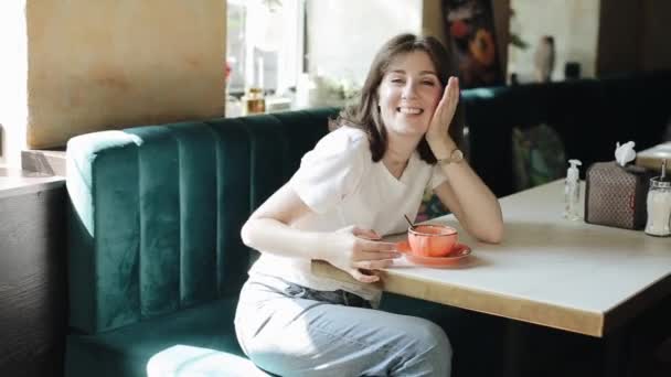 A cheerful young girl in a white T-shirt and jeans sits in a cafe on a summer day and drinks coffee. Front view. Medium plan — 비디오