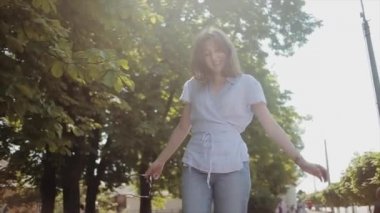 Çekici genç bir kadın ağaçların arka planında güneş ışığında yürür ve gülümseyerek saçlarını düzleştirir. Düşük açı. Ağır çekim. Kamera odağı değiştirir.