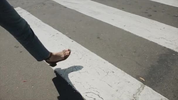 Młoda kobieta w dżinsach i letnich sandałach spaceruje po przejściu dla pieszych skacząc tylko po białych liniach. Zbliżenie nóg. Zwolniony ruch — Wideo stockowe