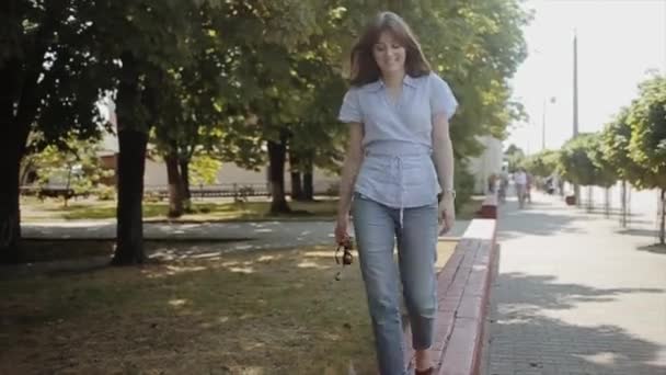 En glad ung kvinna i en ljusblå blus och jeans går längs en tegelsten trottoar med solglasögon i handen och låtsas falla. — Stockvideo