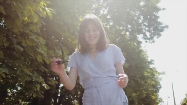 Souriante jeune fille en vêtements d'été légers va sur le fond des arbres verts dans les rayons de lumière diffuse chaude et met des lunettes de soleil. Angle de vue bas. Mouvement lent — Video