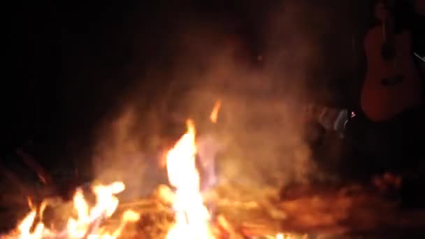 Widok płomieni i dymu z nocnego ogniska na pierwszym planie i człowiek stojący obok niego z gitarą — Wideo stockowe