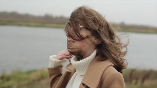 A smiling young girl in a coat stands against the background of the river and straightens her hair, disheveled from the wind. Slow motion — Stock Video