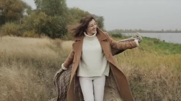 Una joven alegre con gafas recorre el campo en un día de otoño y esconde su cara detrás de una bufanda a cuadros. Movimiento lento — Vídeos de Stock