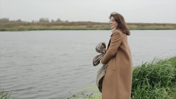 Чарівна молода жінка в коричневому пальто стоїть біля річки і тримає в руках картатий шарф захоплюється природою — стокове відео