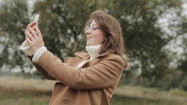 Sonbahar montu giymiş genç bir kız büyük bir ağacın arkasında duruyor ve kameraya gülümserken selfie çekiyor.