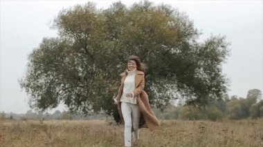 Gülümseyen genç bir kız, rüzgarlı bir sonbahar gününde, nehrin kenarındaki ağaçların arkasında durur ve kendini sıcak bir süvetere sarar.