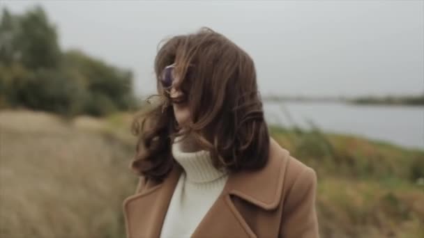 Smyslná mladá žena v brýlích stojí za větrného podzimního dne se zavřenýma očima na břehu řeky a v ústech drží stéblo trávy. Zpomalený pohyb — Stock video