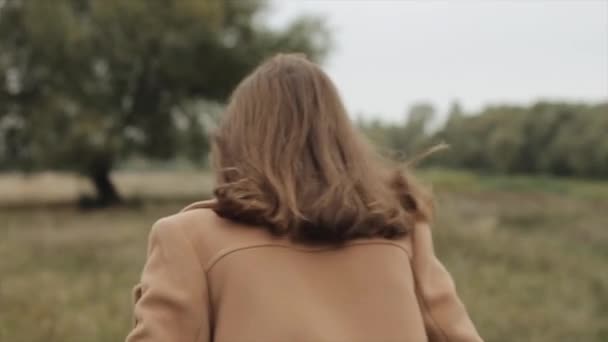 A pleasant young woman in a coat runs across the field to a large tree and turns to look at the camera. Slow motion — Stock Video