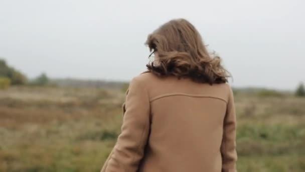 Słodka młoda dziewczyna w okularach przechodzi przez pole w rozpiętym płaszczu i odwraca się do aparatu uśmiechając — Wideo stockowe
