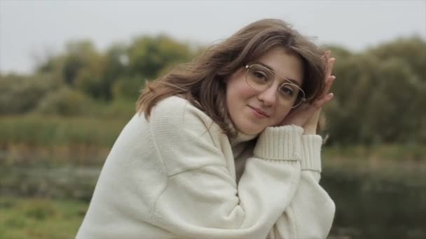 Романтична молода дівчина в білому светрі і окулярах сидить на березі річки і дивиться в камеру, піднімаючи голову на її руках — стокове відео