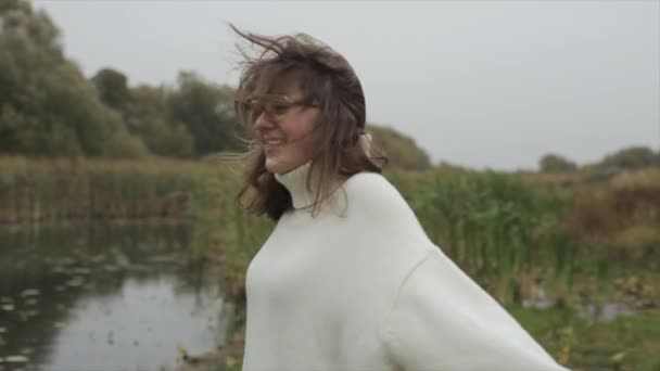 A smiling young girl stands against the background of trees by the river on a windy autumn day and wraps herself in a warm sweater — Stock Video