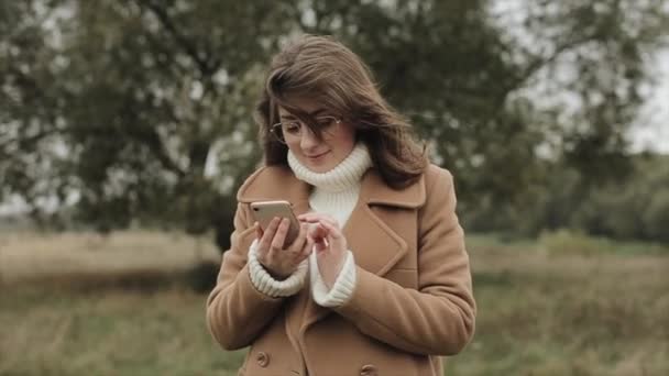 コートを着た穏やかな若い女性が大きな木の背景に立って、携帯電話の画面をクリックします。スローモーション — ストック動画