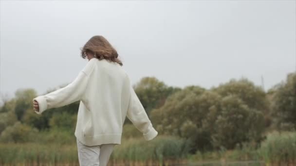 En charmig ung kvinna i en varm vit tröja och glasögon går längs flodstranden och beundrar naturen medan du hukar dig — Stockvideo