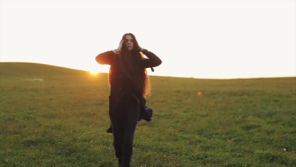 Chica joven sensual en ropa negra va a lo largo de las verdes colinas contra la puesta de sol y mira a la cámara. Movimiento lento — Vídeo de stock