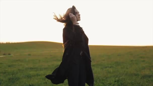Ніжна молода дівчина йде вздовж зеленого лугу і кружляє проти заходу сонця, випрямляючи її довге волосся. Повільний рух — стокове відео