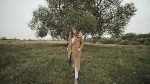 Cheerful young girl in glasses walks across the field in a brown coat wide open and straightens her hair tousled by the wind — Stock Video