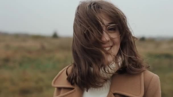 Eine sanfte junge Frau steht im Herbstmantel vor dem Hintergrund der Natur und korrigiert ihre im Wind zerzausten Haare. — Stockvideo