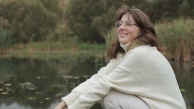 Beyaz elbiseli, gözlüklü hoş bir kız nehre çömelip suya bakıyor.