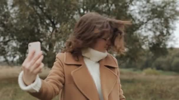 Glada ung kvinna i glasögon står i en rock på en bakgrund av ett träd och lurar runt tittar i mobiltelefon skärm — Stockvideo