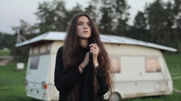 검은 옷을 입은 한 젊은 머리의 소녀가 여행 트레일러 뒤의 푸른 초원에 서서 주위를 둘러 본다. 느린 동작 — 비디오